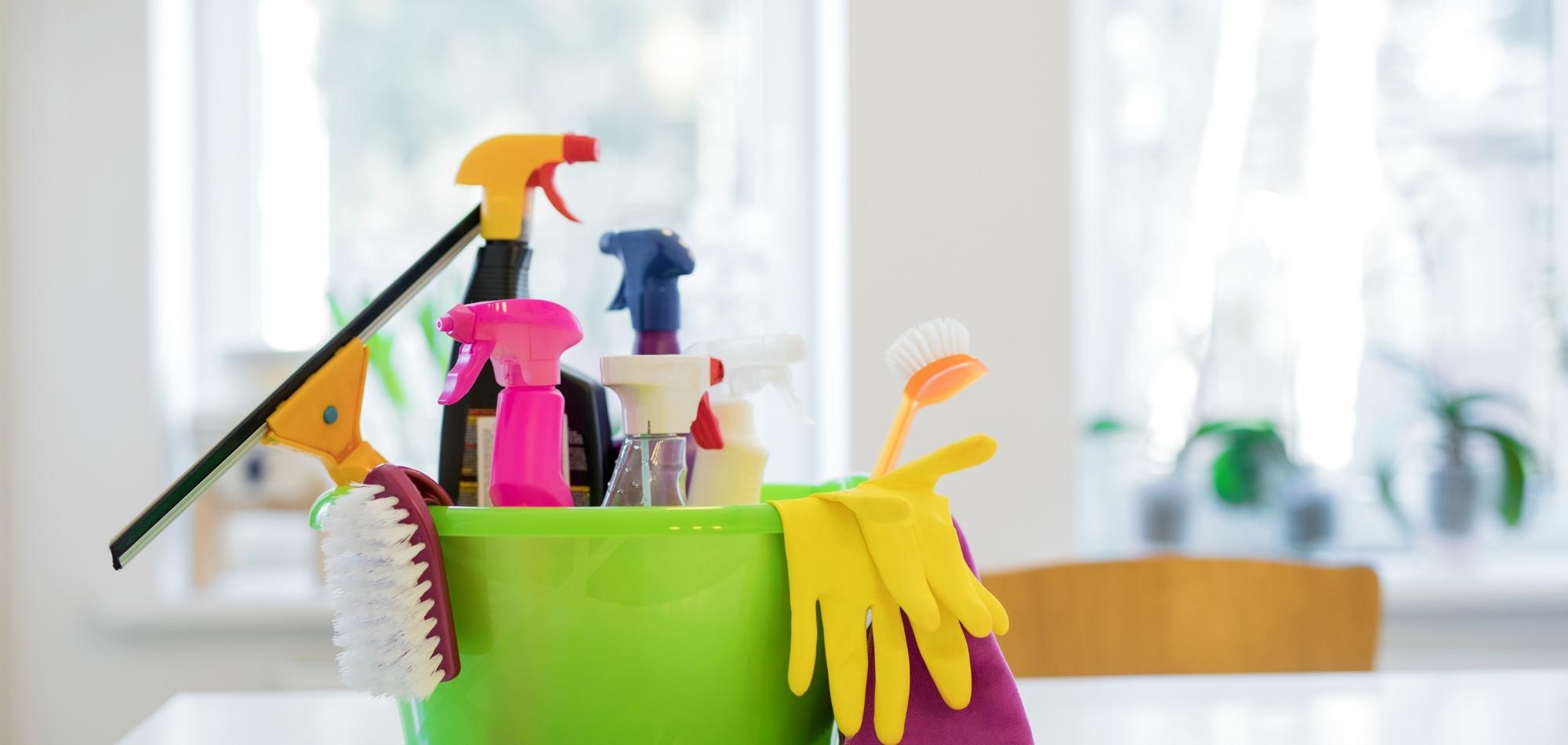 A green bucket filled with various cleaning supplies, perfect for domestic cleaning, sits on a white surface with a blurred background of a bright room featuring windows and plants.