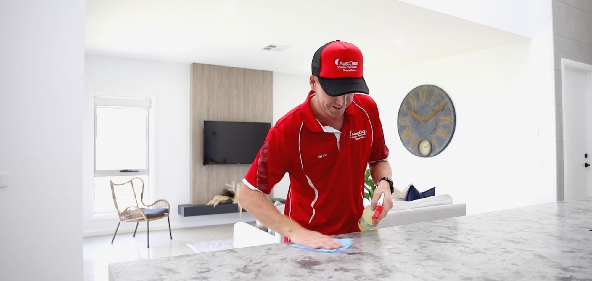 A person wearing a red shirt and cap is busy with spring cleaning, wiping down a marble worktop in a modern, bright living room featuring a wall-mounted TV and a large round clock in the background.