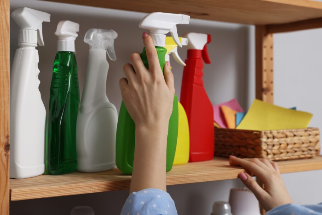 A person reaches for a green spray bottle among various coloured home cleaning bottles on a wooden shelf, with a basket of cleaning cloths nearby.