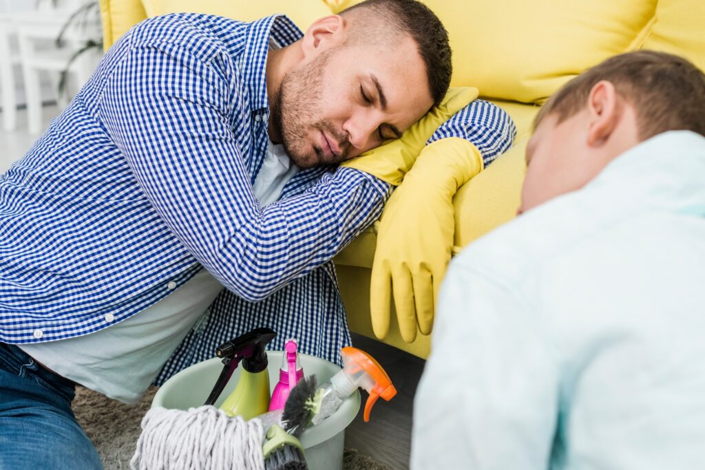 A man wearing yellow cleaning gloves is sleeping on a sofa, resting his head on his arm after a long day of house cleaning, with a cleaning caddy and mop nearby. Another person is also visible, facing away and looking tired.