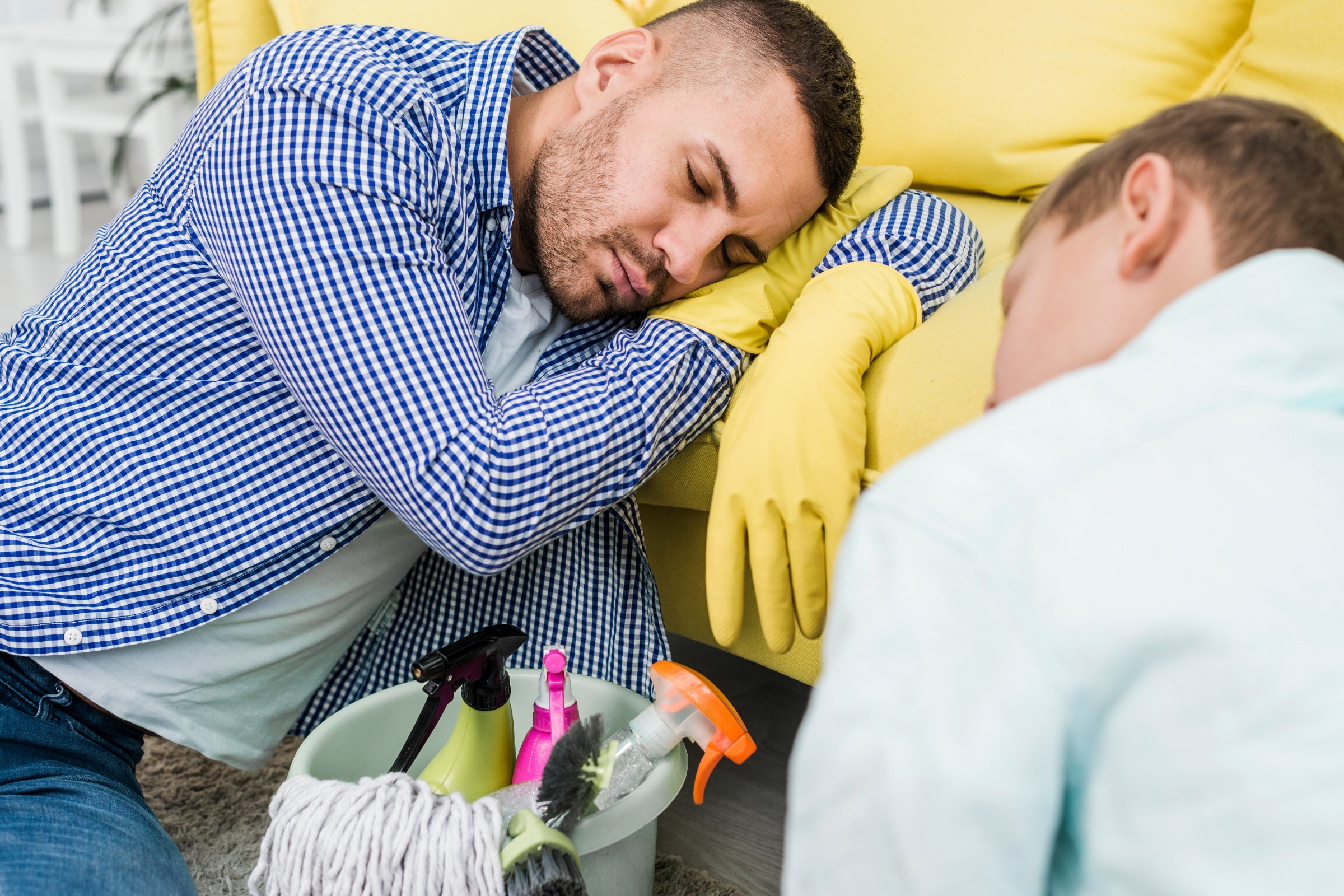 A man wearing yellow cleaning gloves is sleeping on a sofa, resting his head on his arm after a long day of house cleaning, with a cleaning caddy and mop nearby. Another person is also visible, facing away and looking tired.