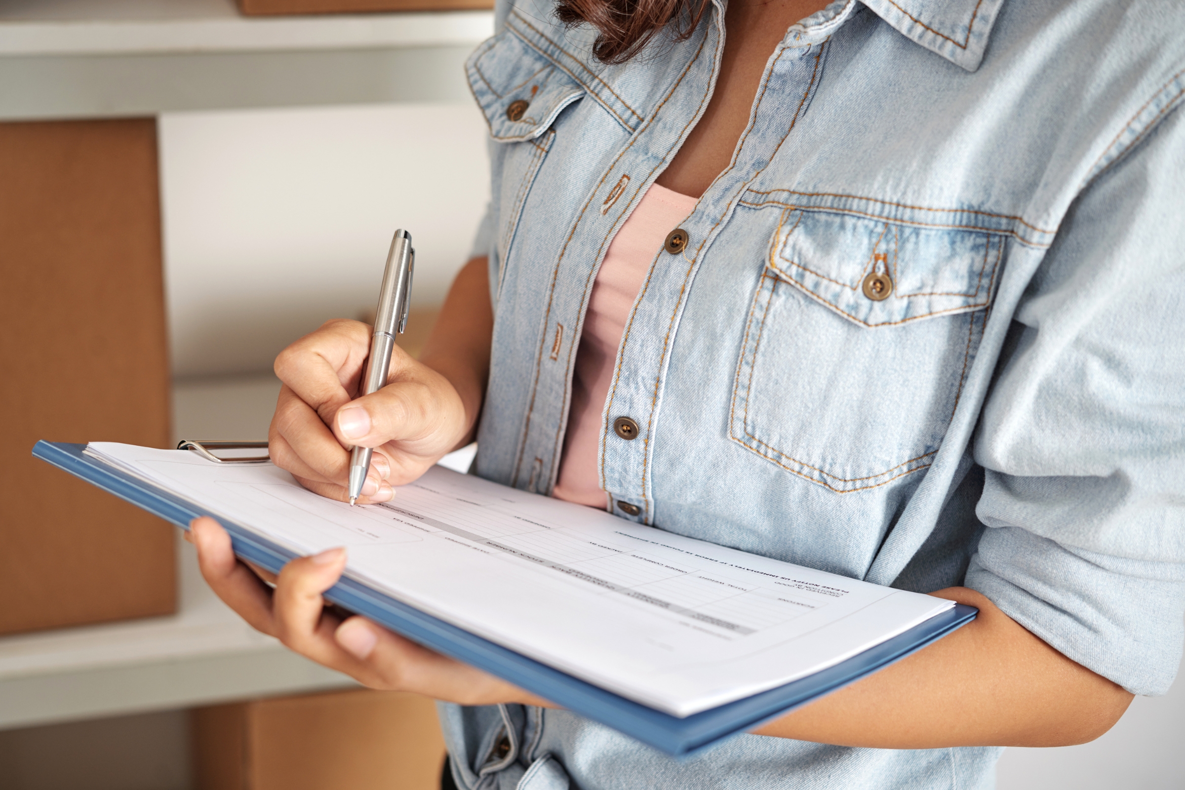 A person in a denim shirt is holding a clipboard and writing on a form, possibly organising home cleaning supplies, with cardboard boxes on shelves in the background.