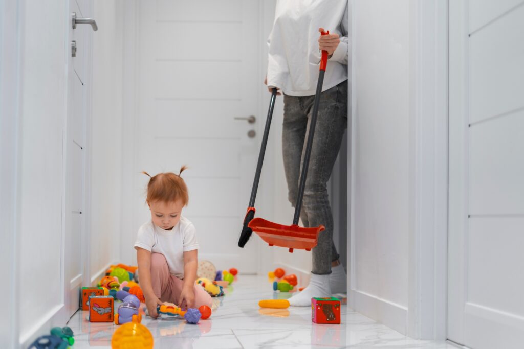 A toddler sits on a hallway floor surrounded by colourful toys, while an adult stands nearby holding a broom and dustpan, ready for some house cleaning.