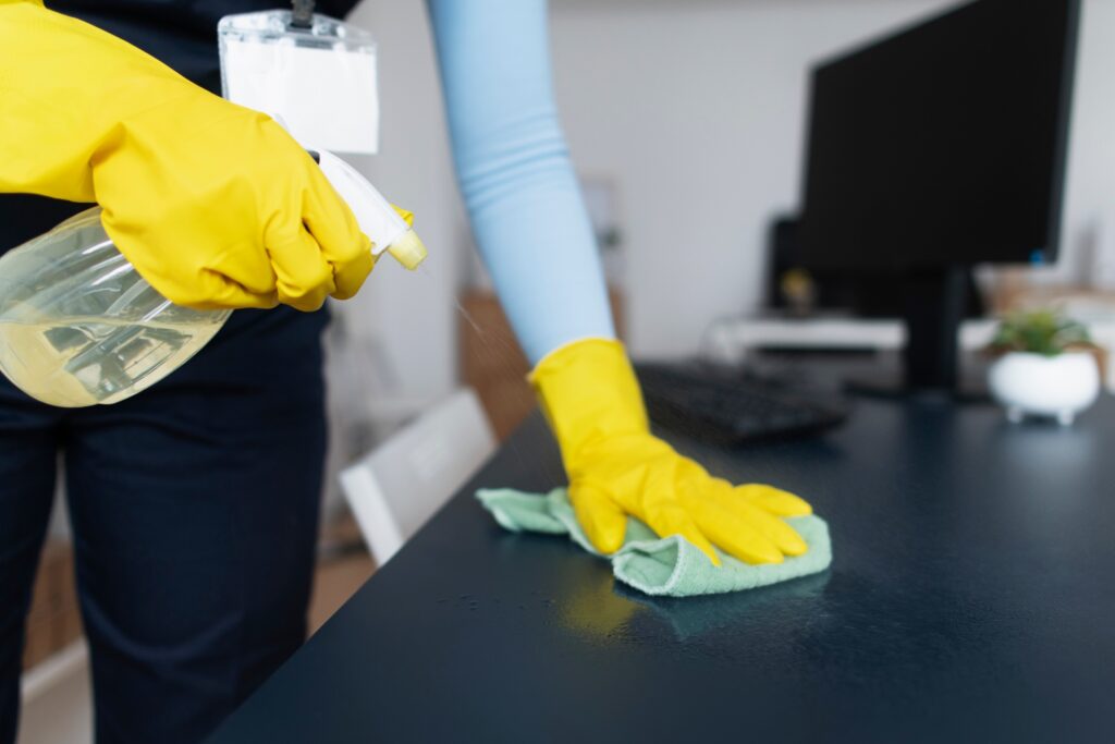 A person wearing yellow rubber gloves cleans a dark desk with a green cloth and spray bottle in an office setting; a computer monitor, keyboard, and AustClean's commitment to spotless workspaces are visible in the background.