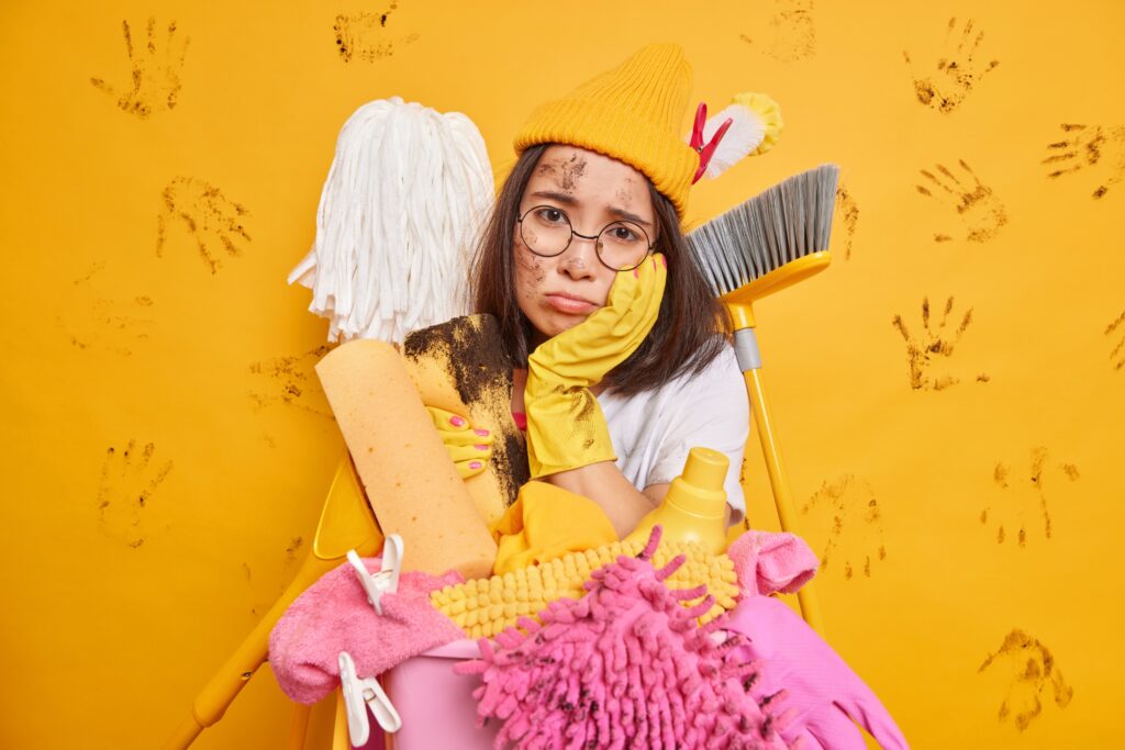 A tired woman in cleaning gloves and a yellow hat, with a dirty face, holds household cleaning supplies against a yellow wall marked by muddy handprints. She looks frustrated and overwhelmed.