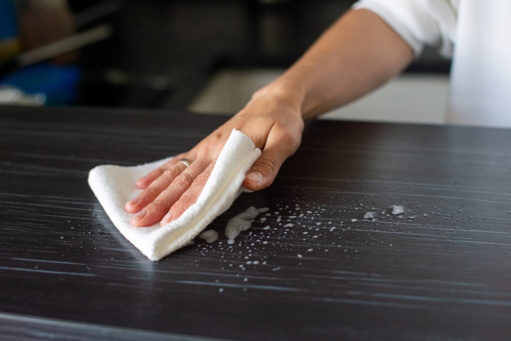 A person wipes a dark worktop with a white cloth, cleaning up soapy water to reduce dust. Only their hand and part of their arm are visible.